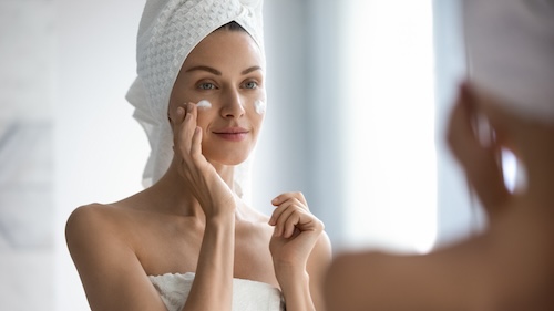 Aantrekkelijke jonge volwassen vrouw die gezichtscrème op gezicht doet voor een glowy look