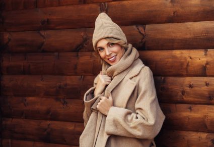 Modekleuren Herfst: vrouw met gamel kleurige jas en muts op, poseert voor een houten schutting