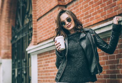 Leren Jassen: Jonge en mooie moderne vrouw in de stad, staand op de stoep met een kop koffie in haar hand en een leren jas aan.