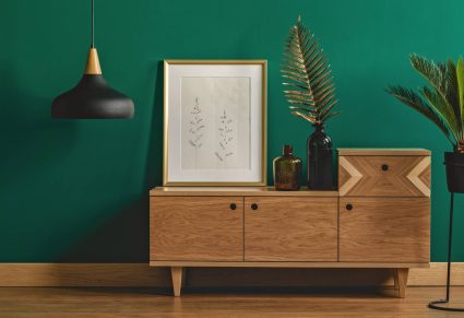 Goedkope Accessoires Xenos: Industriële hanglamp naast een stijlvol dressoir en een kunstposter in een gouden frame bij een donkergroene muur van een modern interieur