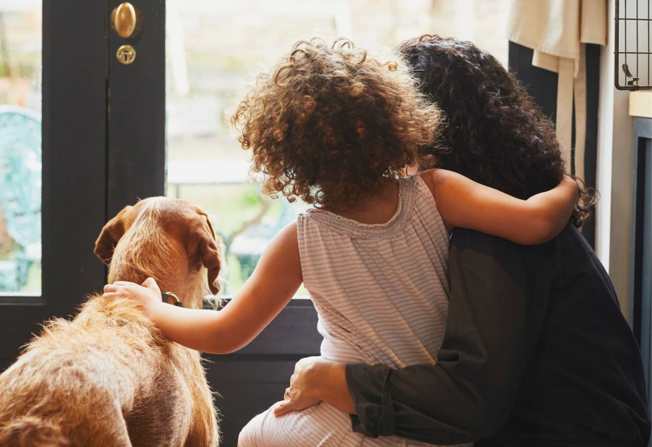 Hond Deel Van Het Gezin: Moeder en jonge dochter kijken uit het keukenraam met de hond van de familie. Dochter zit op moeders schoot met armen in een omhelzing, meisje aait hond.