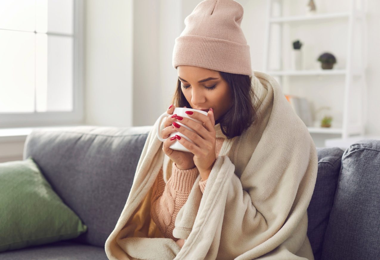 Plaids: Jonge vrouw met gebreide trui en muts zit thuis op de bank in een plaid warme koffie of thee te drinken.